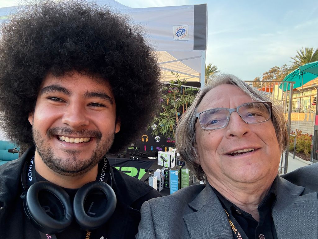 Two men smiling for a selfie at an outdoor event. One has an afro hairstyle and headphones around his neck; the other has gray hair and glasses. Booths and plants are visible in the background under a clear sky.
