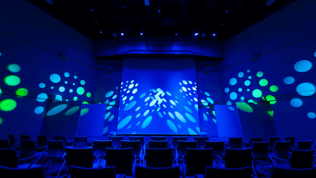Rows of empty chairs face a stage with blue lighting and green and white spot patterns projected on the walls, creating a vibrant, atmospheric setting in a modern auditorium.