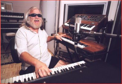 A man with long hair and sunglasses smiles while playing a keyboard in a recording studio, surrounded by synthesizers and a microphone.