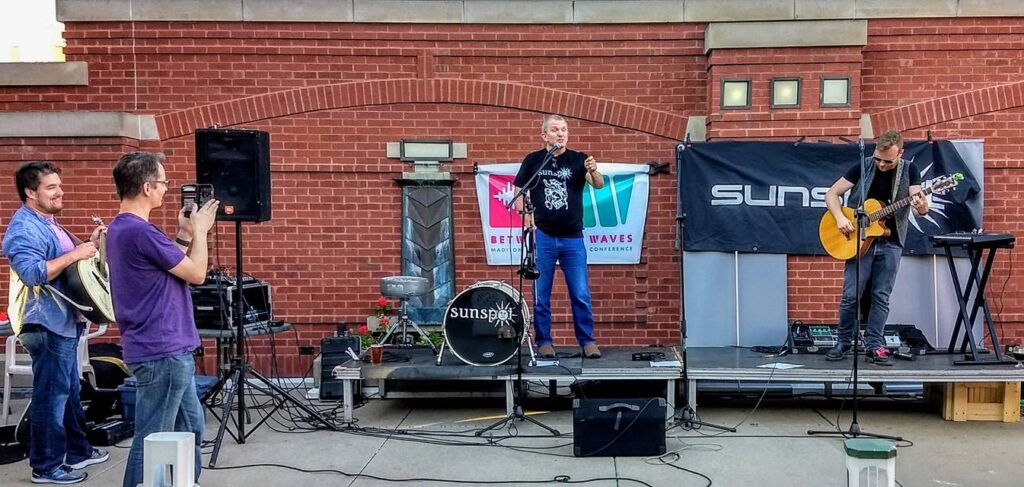 A small band performs on an outdoor stage with brick walls behind them. One person sings, another plays guitar, and a third plays keyboard. Two people offstage watch and take photos. Banners hang on the wall behind the stage.