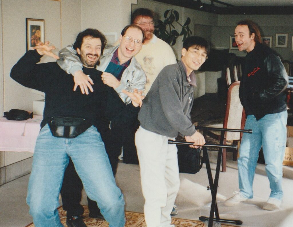 Five men pose playfully indoors, with one man pretending to grab another from behind. They are all smiling or laughing, dressed casually, and standing near a table and chairs in a warmly lit room.