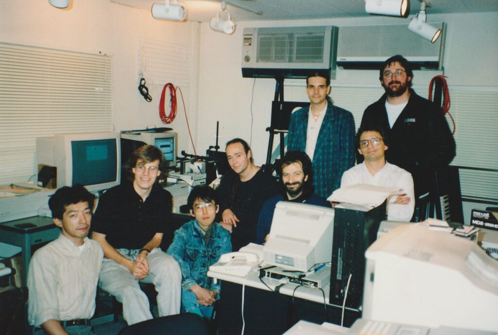 Eight people pose together in a cluttered office filled with computers, cables, and monitors. Some are seated in front of the equipment, while others stand behind them, all smiling for the camera.