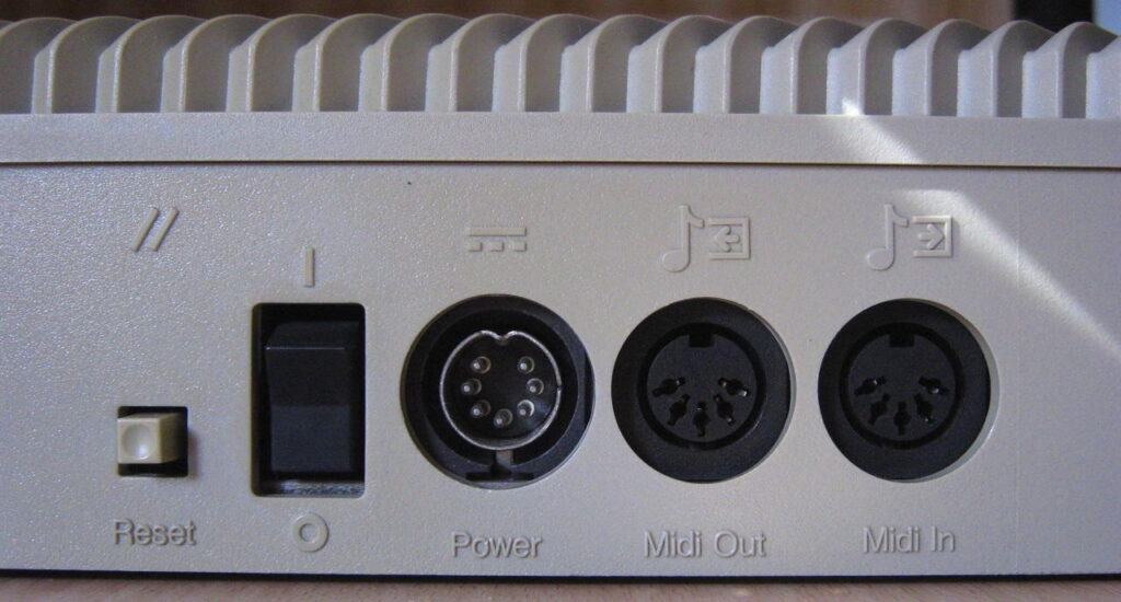 Close-up of the back panel of a vintage computer or synthesizer, showing a reset button, power switch, round power connector, and MIDI Out and MIDI In ports.