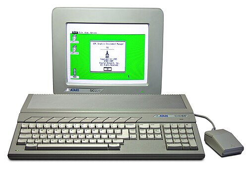 An Atari 1040ST personal computer with a monitor displaying a green-background desktop interface and a mouse connected on the right side.