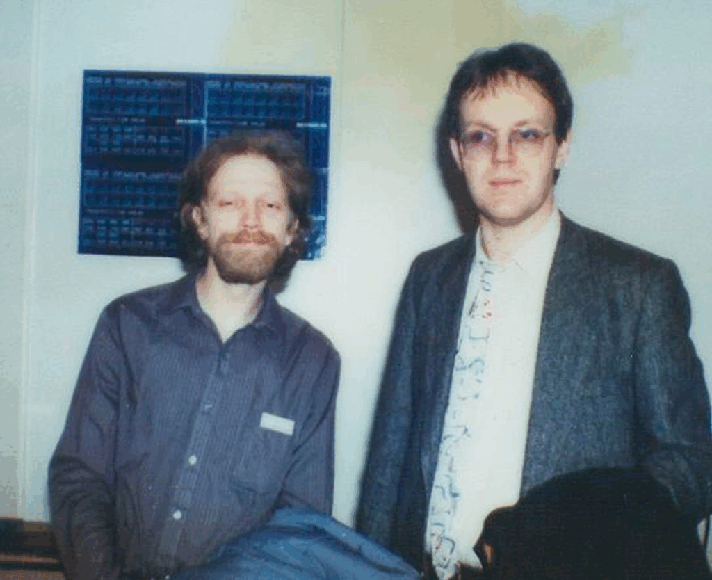 Charlie Steinberg and Manfred Rurup standing indoors, one with a beard and long hair, wearing a striped shirt, the other with short hair, glasses, a suit jacket, and a patterned tie. A blue electronics panel is mounted on the wall behind them.