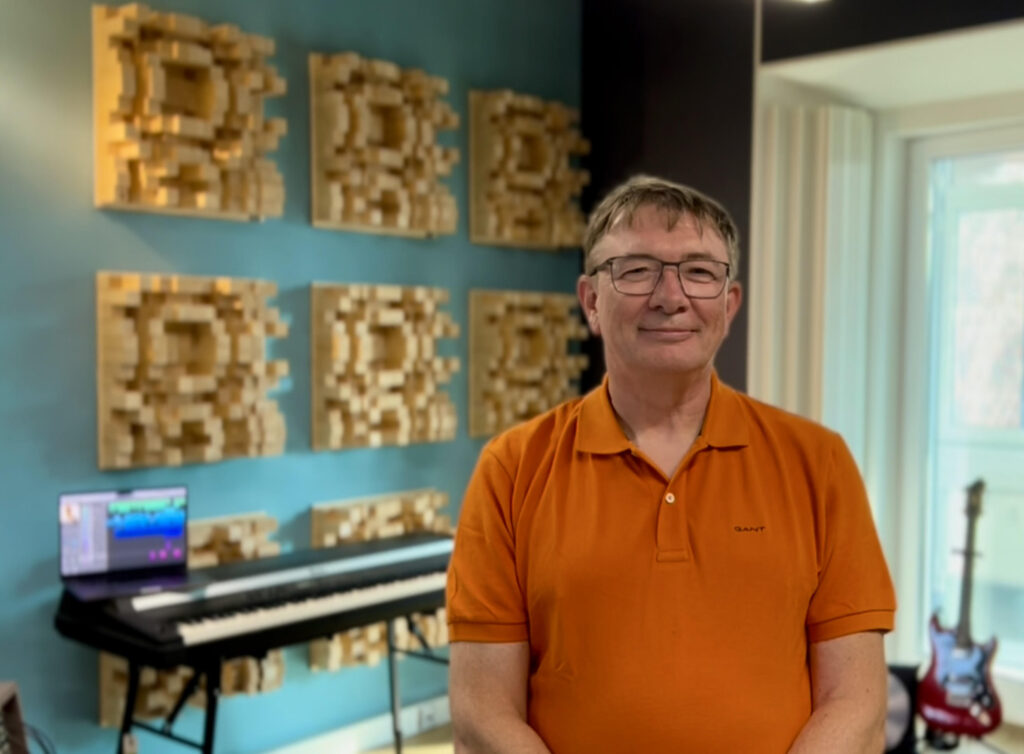 A man in an orange polo shirt stands smiling in a music studio with a keyboard, a laptop, a guitar, and decorative acoustic panels on a blue wall behind him.