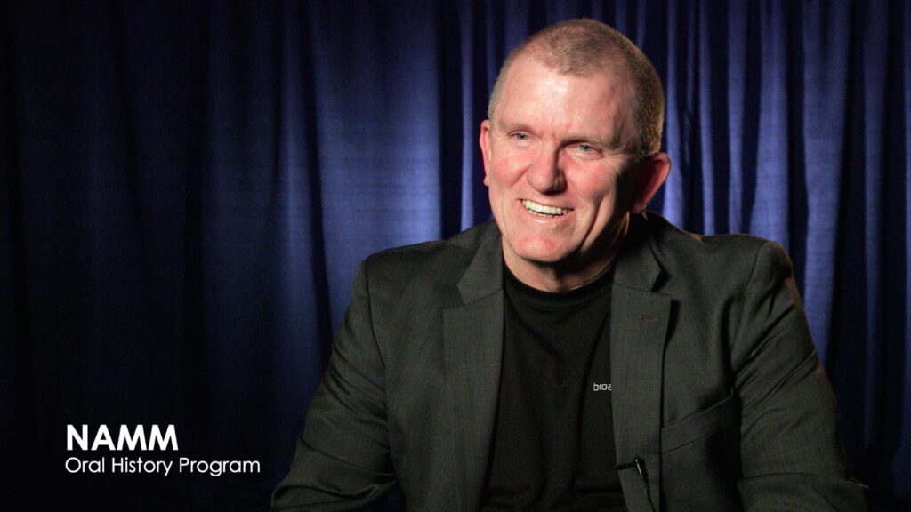 A man in a dark blazer and black shirt smiles while seated in front of dark blue curtains. White text in the lower left reads, NAMM Oral History Program.