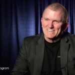 A man in a dark blazer and black shirt smiles while seated in front of dark blue curtains. White text in the lower left reads, NAMM Oral History Program.