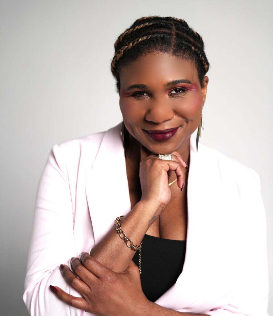 A woman with braided hair, wearing a light pink blazer over a black top, smiles with one hand resting under her chin and the other arm folded. She has bold makeup and wears a bracelet and ring, posing against a plain background.