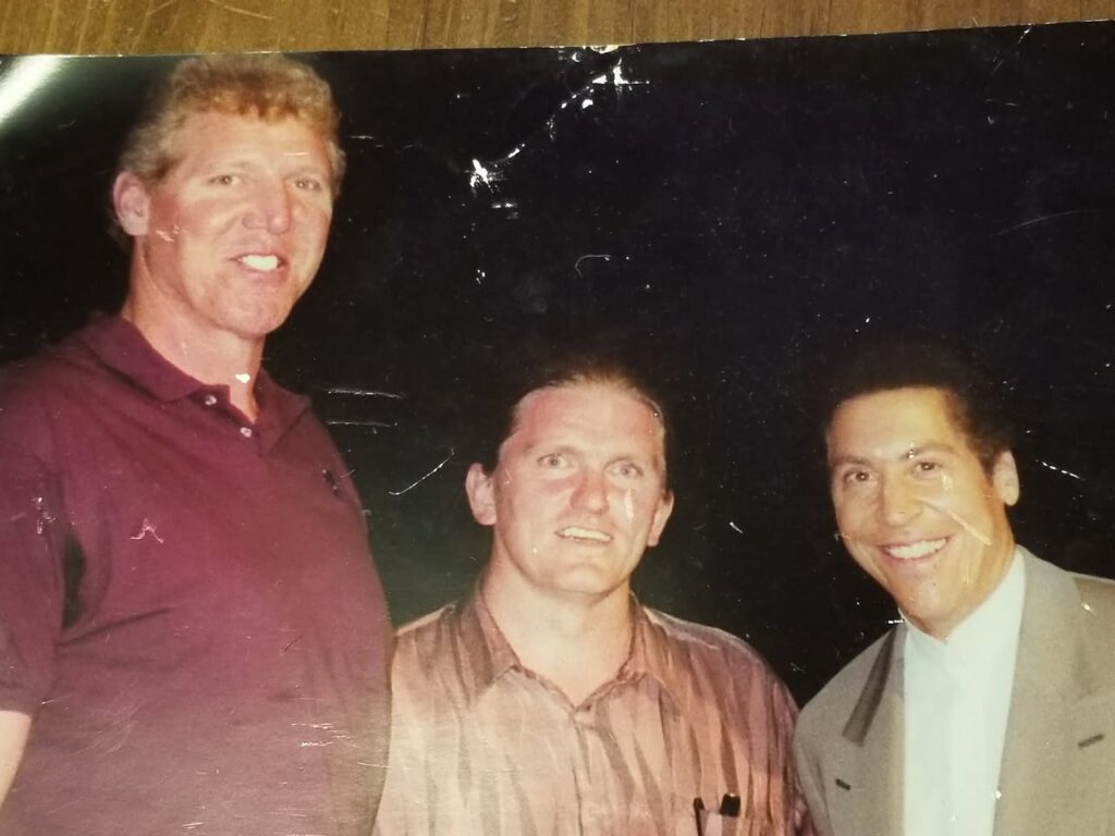 Three men are posing together and smiling for a photo. The man on the left is tall and wearing a maroon polo shirt, the man in the middle has a striped shirt, and the man on the right is in a light-colored suit against a dark background.