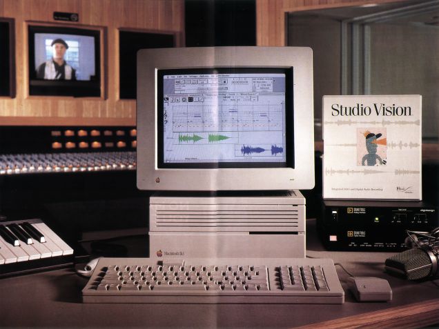 A vintage Macintosh computer displays audio editing software in a recording studio, surrounded by a keyboard, MIDI controller, Studio Vision manual, and professional audio equipment.