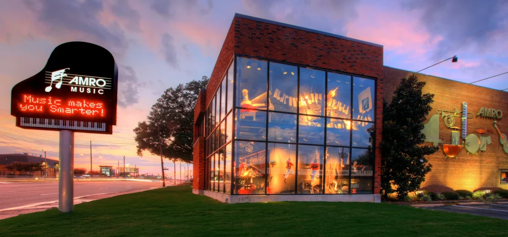 A modern brick building with large glass windows displays musical instruments inside. A sign shaped like a grand piano reads AMRO MUSIC and Music makes you Smarter! at sunset.