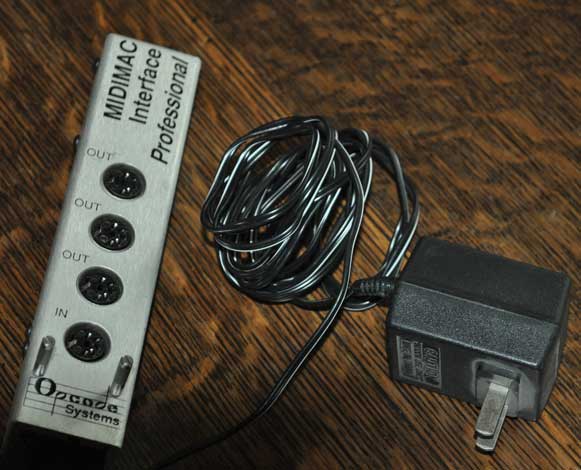 A MIDI interface device labeled MIDIMAC Interface Professional with four round MIDI ports and a black power adapter with a coiled cord, placed on a wooden surface.
