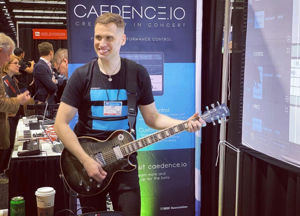 A man plays an electric guitar at a booth for Caedence.io at a convention. He smiles while wearing a badge and a branded shirt. People and electronic equipment are visible in the background.