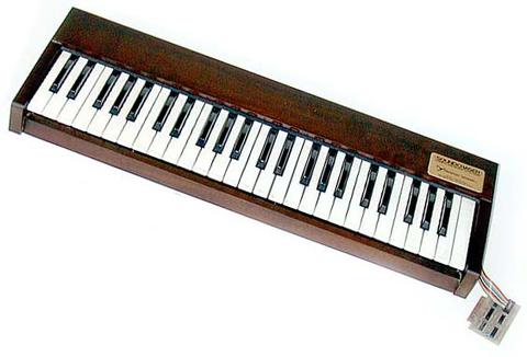 A vintage wooden electronic keyboard instrument with black and white keys and a small control panel on the top right corner, connected to a cable.
