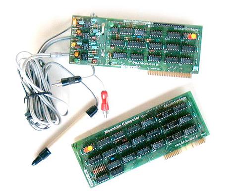 Two vintage computer circuit boards labeled Mountain Computer Music System are shown, along with a connected gray cable ending in a black audio jack and a red adapter.