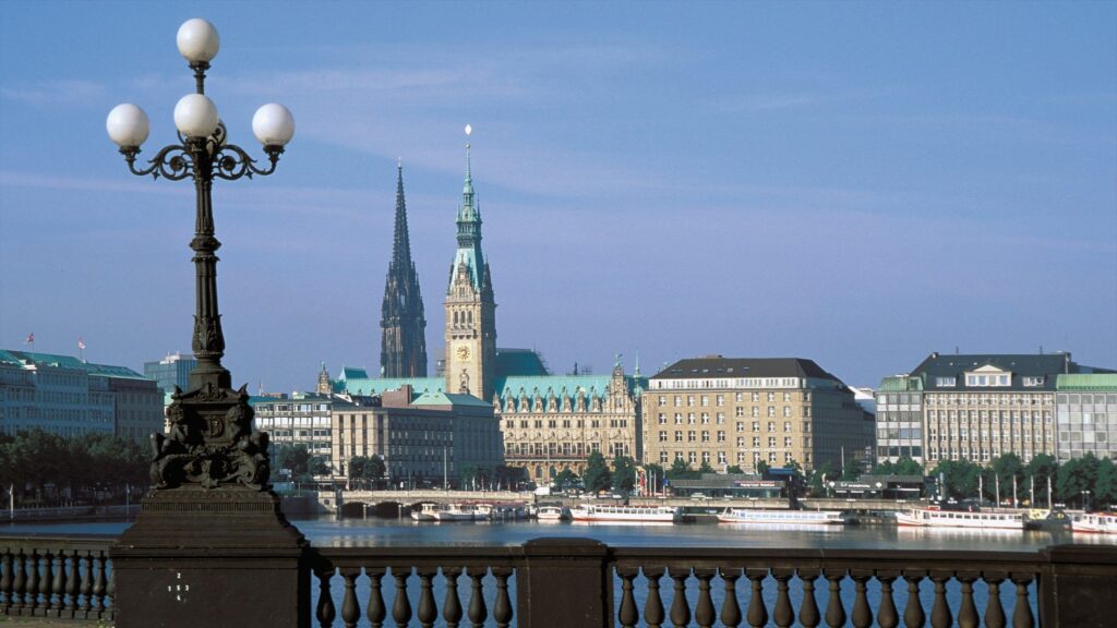 A view of historic buildings and church spires across a body of water in Hamburg, Germany, with a vintage streetlamp and railing in the foreground under a clear blue sky.