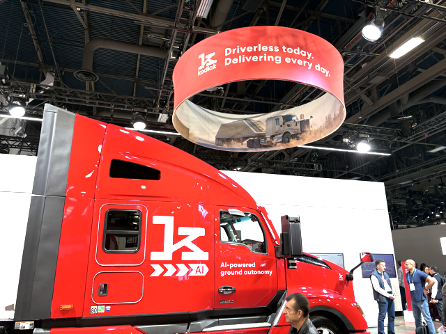 A bright red semi-truck with 1K AI-powered ground autonomy on the side is displayed indoors. Above, a circular banner reads, Driverless today. Delivering every day. Several people stand nearby.
