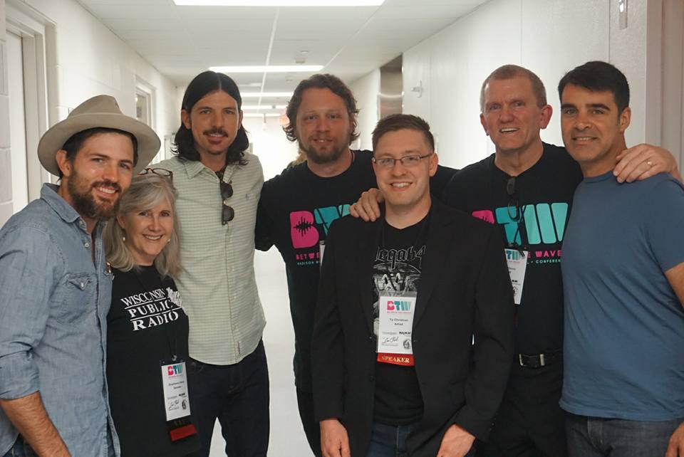 A group of seven adults, including five men and two women, smile and pose together in a brightly lit hallway. Some wear conference name tags and casual clothes, while one woman wears a public radio T-shirt.