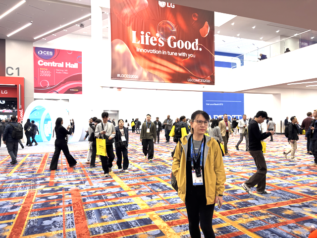 People walk around a large, brightly lit convention hall with colorful carpet. A woman in a yellow jacket stands in the foreground. A big red LG sign above reads, “Life’s Good. Innovation in tune with you.”.
