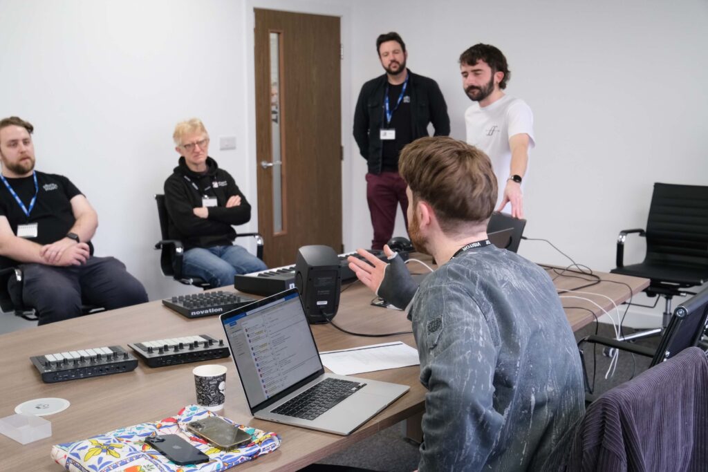 Five people are having a discussion in a meeting room. One person is standing and speaking, while the others are seated around a table with laptops, notebooks, and electronic equipment.