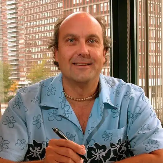 A smiling man in a light blue, flower-patterned shirt holds a pen and sits indoors by large windows, with tall buildings and trees visible outside.