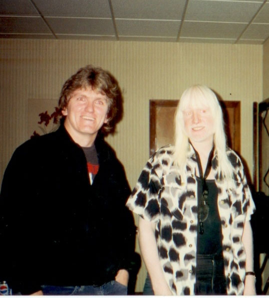 Two people stand indoors, smiling at the camera. The person on the left has medium brown hair and wears a dark jacket, while the person on the right has long, straight, light blonde hair and wears a patterned shirt over black clothing.