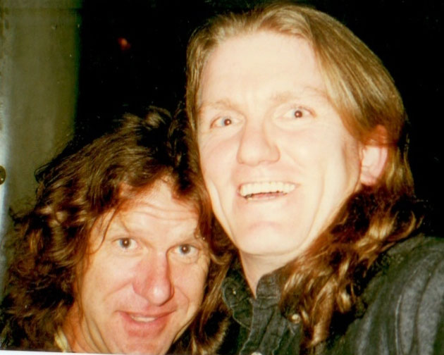 Two men with long brown hair are close together, smiling at the camera in a casual, warmly lit setting.