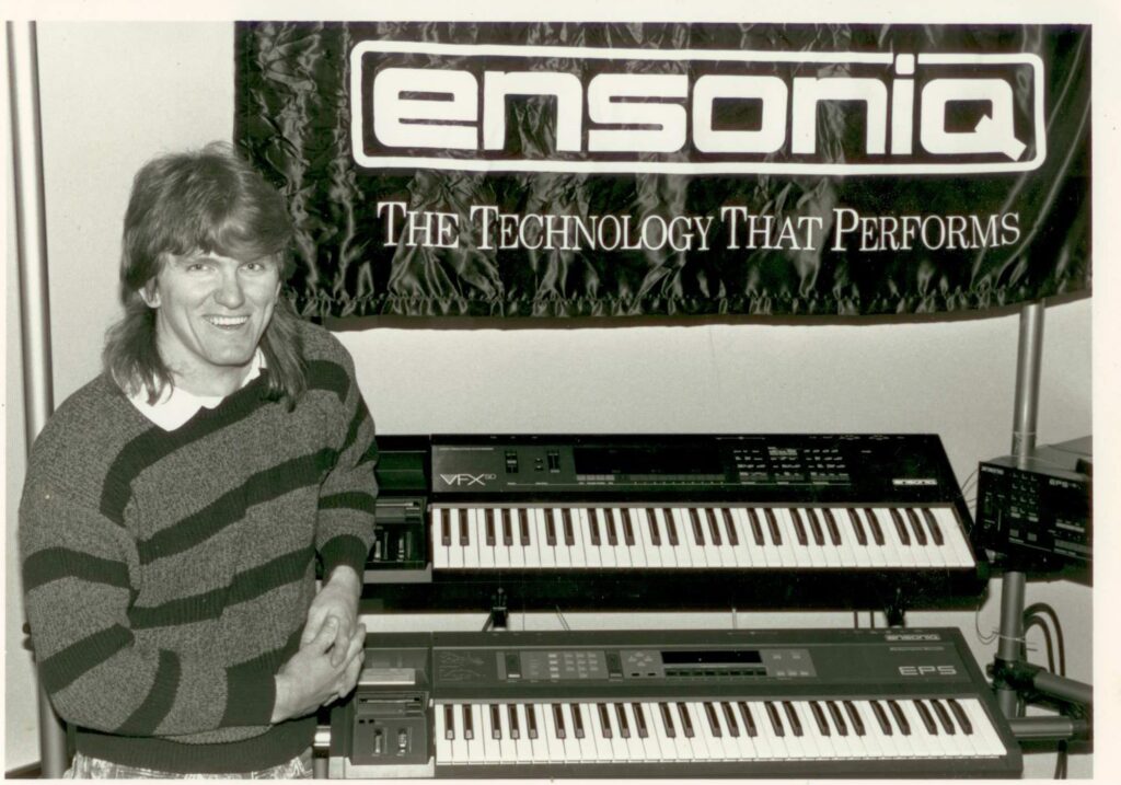 A smiling person with long hair stands in front of two Ensoniq synthesizer keyboards, beneath a banner that reads “Ensoniq: The Technology That Performs.”.