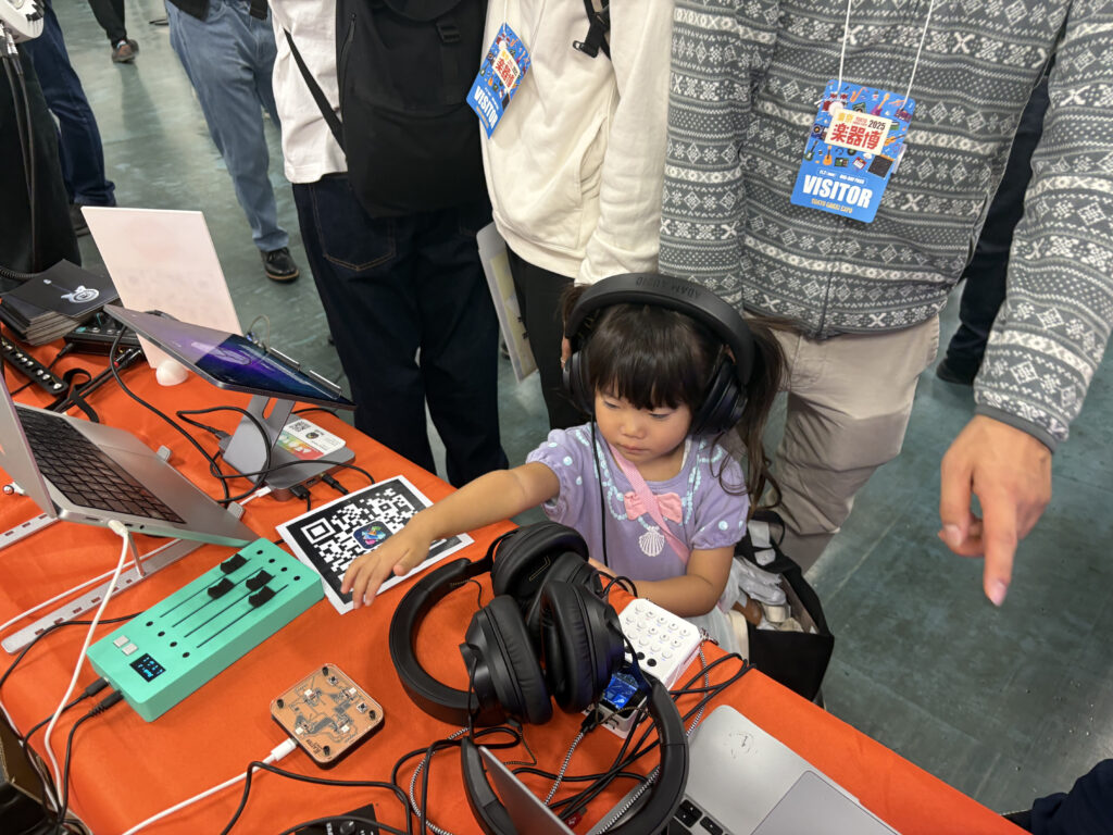 A young girl wearing large headphones interacts with music equipment on a table at an event. Laptops, headphones, and audio devices are scattered across the table. Adults stand nearby, one pointing toward the setup.