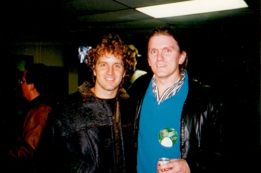 Two men stand close together and smile at the camera in a dimly lit indoor setting. One wears a dark jacket with a fur collar; the other wears a leather jacket and a large green VIP badge. A few people are visible in the background.
