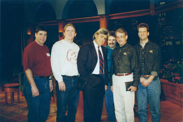 Six men stand together indoors, some smiling, with one dressed in a suit and tie while the others wear casual shirts and jeans or khakis. The background suggests a late-night talk show set.