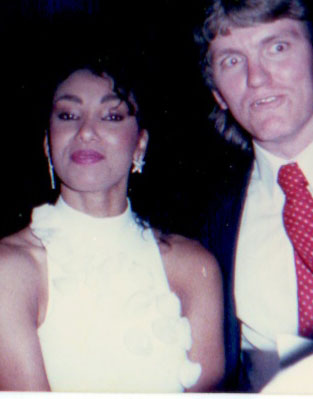 A woman in a sleeveless white dress with floral details stands next to a man in a suit and red tie. Both are facing the camera; the background is dark.