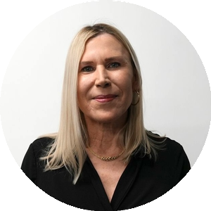 A woman with straight, shoulder-length blonde hair wearing a black top, gold hoop earrings, and a gold necklace, smiling in front of a plain white background.
