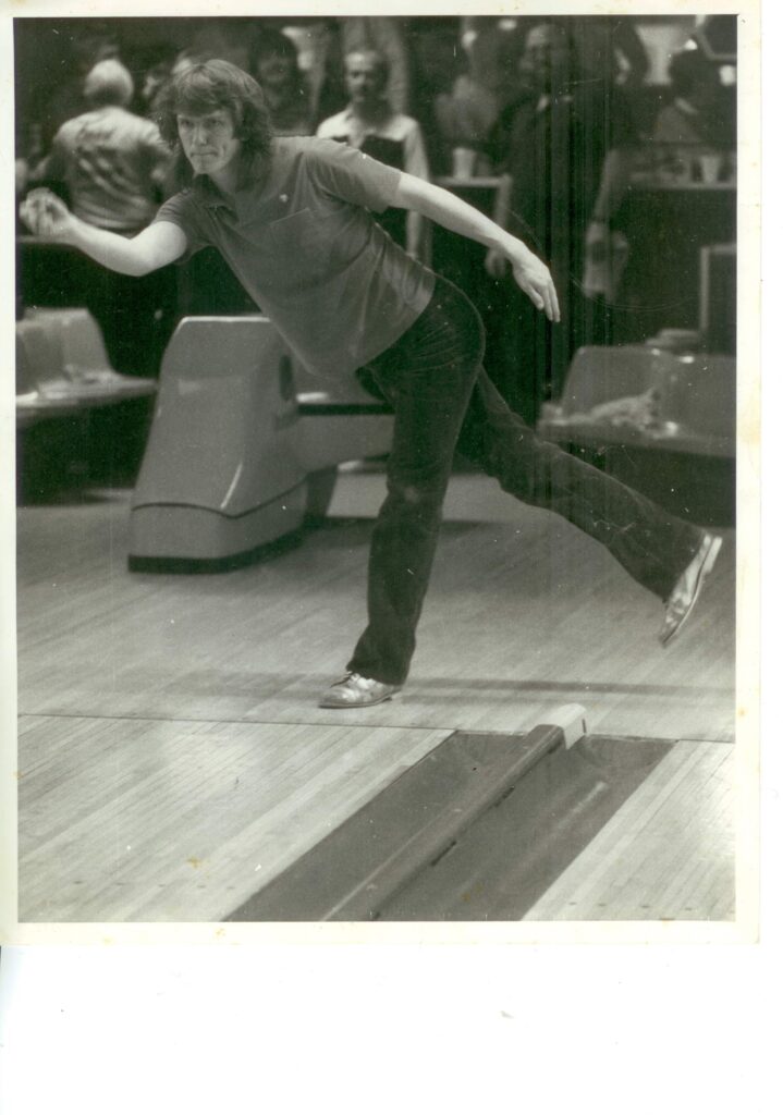 A woman in mid-action bowls a ball down a lane in a bowling alley. She is wearing glasses, a short-sleeve shirt, and dark pants. Several people are visible watching in the background. The image is black and white.