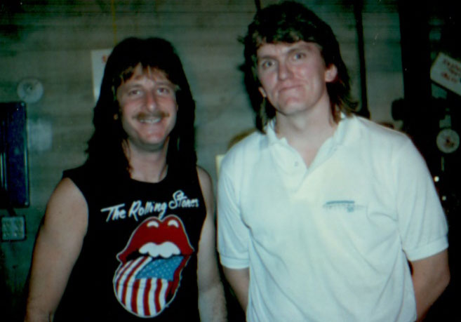Two men pose indoors. One wears a black Rolling Stones sleeveless shirt and has a mullet and mustache, smiling. The other, in a white polo shirt with short brown hair, stands beside him, unsmiling.