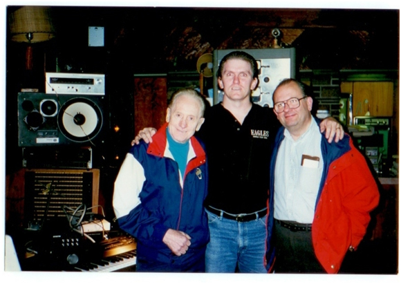 Three men pose together, smiling, in a room with audio equipment, speakers, and a keyboard. Two wear jackets with blue and red colors; the man in the middle wears a black shirt and jeans. The background is dimly lit.