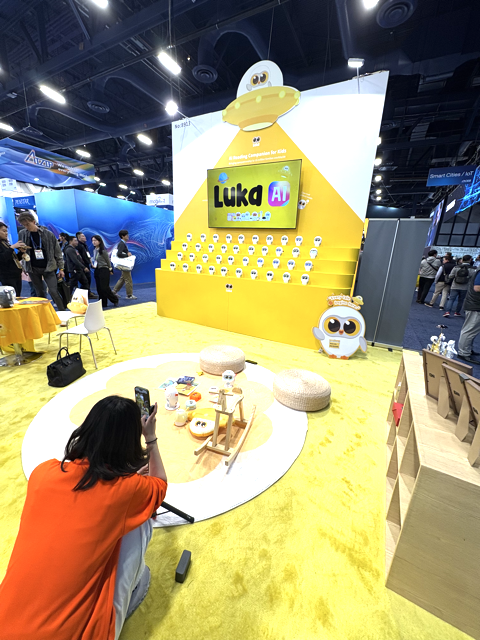 A person in an orange shirt takes a photo of a yellow and white Luka AI display at a convention, featuring toy mascots, a large screen, and a play area with stools and children’s toys. People walk in the background.