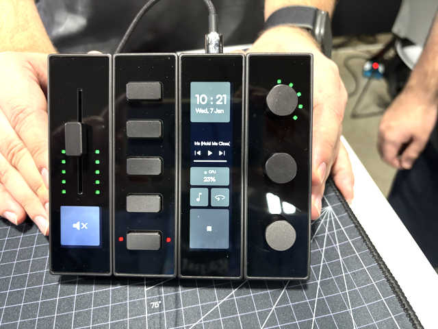 A person holds a modular audio control device with five sections, including sliders, buttons, dials, and a central digital display showing the time, date, battery level, and media controls.