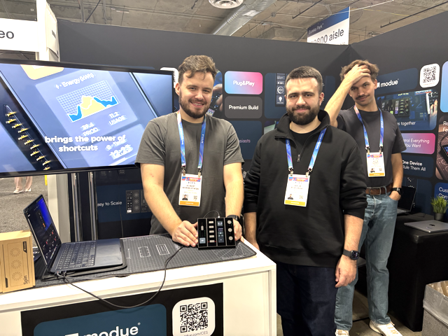Three men stand at a tech booth. Two smile at the camera, one resting his hand on a keypad device. The third man stands behind, touching his head. A monitor and product display are visible at the booth.