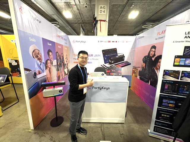 A man stands smiling and gesturing toward a display booth for PartyKeys, featuring colorful banners, electronic keyboards, and promotional materials at an indoor event or convention.