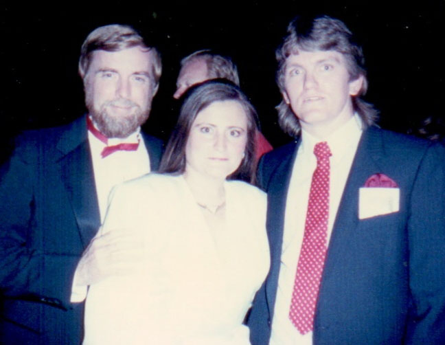 Three people in formal attire pose together at an event. The man on the left has a beard and wears a black suit with a red bow tie. The woman in the center wears white. The man on the right wears a suit with a red polka dot tie.