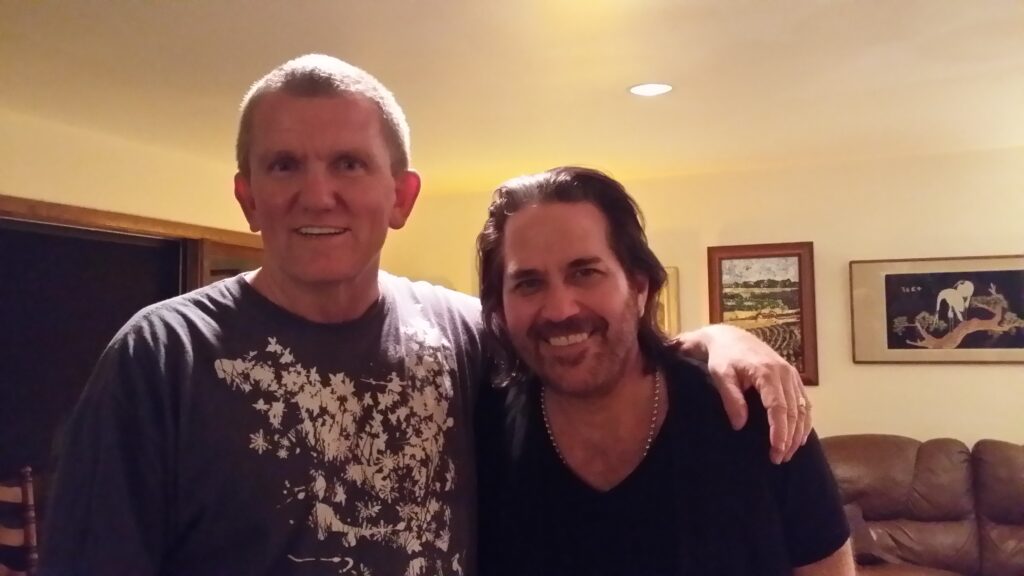 Two men are standing close together indoors, smiling at the camera. One man has his arm around the other. There are paintings and a couch visible in the background.