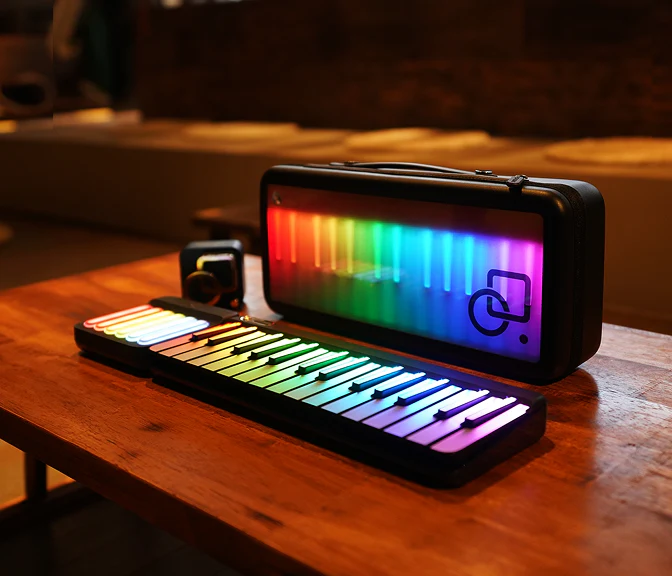 A colorful, illuminated electronic keyboard and matching carrying case sit on a wooden table in a cozy indoor setting. The keys and case both display a vibrant rainbow gradient.