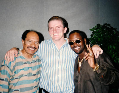 Three men stand close together smiling at the camera. The man on the left wears a striped shirt, the man in the center wears a striped button-down shirt, and the man on the right wears sunglasses and a dark shirt, holding up one finger.