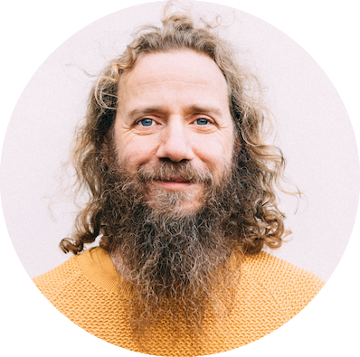 A man with curly, long hair and a full beard smiles gently. He is wearing a textured mustard-yellow sweater and standing against a plain, light-colored background.