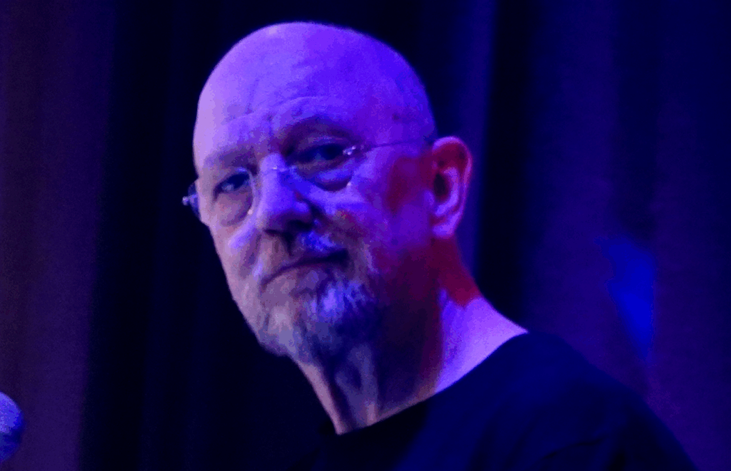 A bald man with glasses and a gray beard, wearing a black shirt, is illuminated by blue and purple lighting, standing in front of a dark, blurred background.