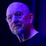 A bald man with glasses and a gray beard, wearing a black shirt, is illuminated by blue and purple lighting, standing in front of a dark, blurred background.