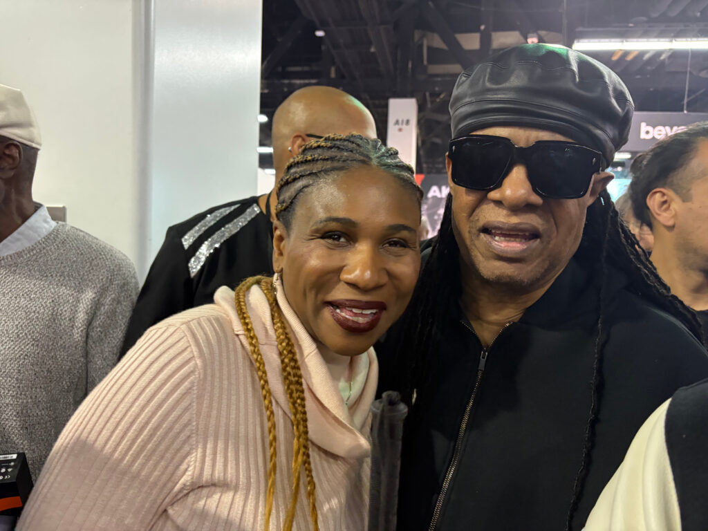 A woman with braided hair and a light pink sweater smiles and poses next to a man wearing sunglasses, a black leather cap, and a black jacket in a crowded indoor setting.