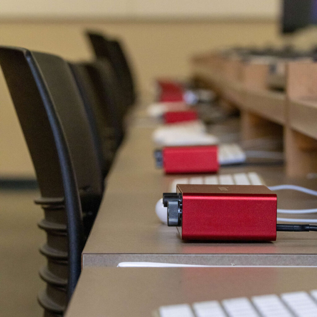 Several red electronic devices connected to white cables are arranged in a row on a long desk in a computer lab, with black chairs lined up beside the desk.